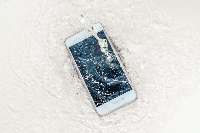 Smartphone submerged, water splashing vigorously, creating ripples across a smooth white surface.