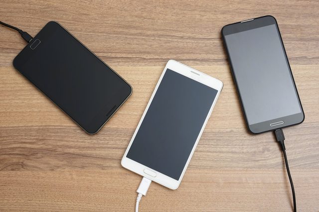 Three smartphones, connected to chargers, rest on a wooden surface.