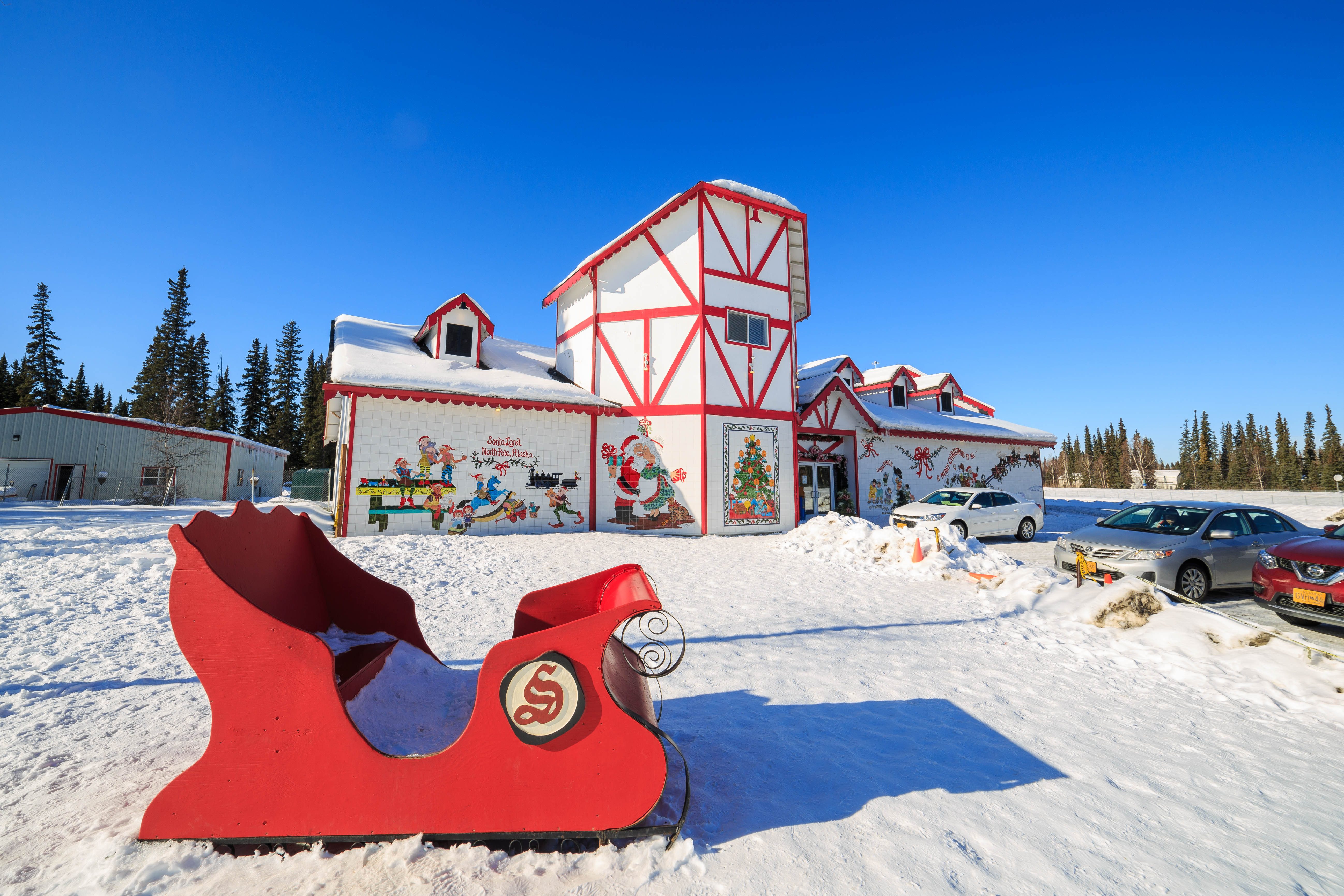 Red sleigh sits on snowy ground; festive building features Christmas murals and trees, surrounded by parked cars and distant evergreens under a clear blue sky.