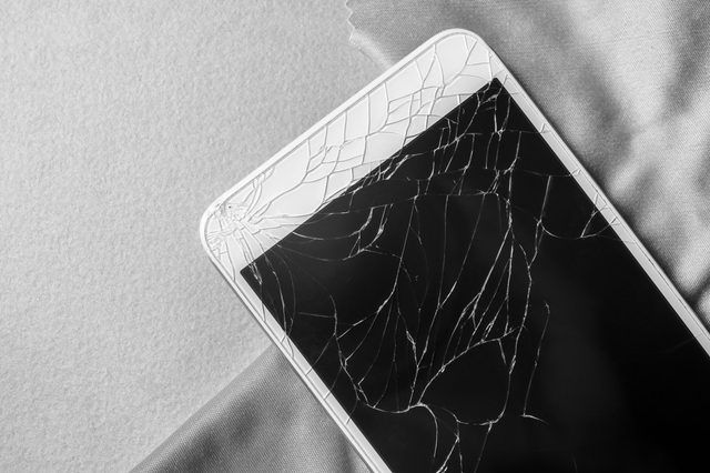 A cracked smartphone screen rests on textured fabric, displaying intricate spiderweb-like fracture patterns in grayscale.