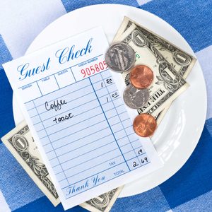 Guest check listing coffee and toast with prices on a white plate alongside dollar bills and coins, placed on a blue checkered tablecloth.