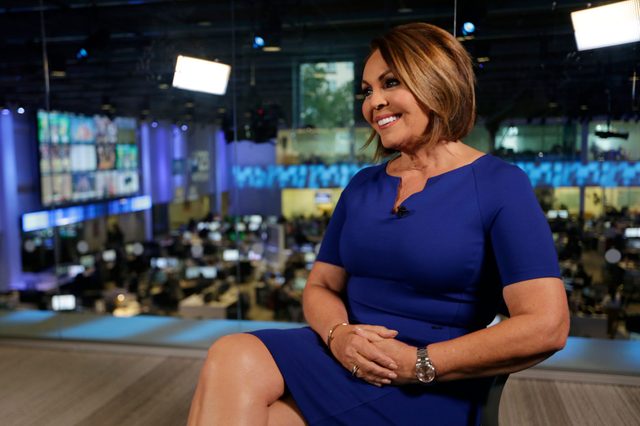 Person smiling, sitting in a blue dress, in a busy newsroom with screens and computers in the background.