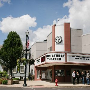 Theater marquee displays show dates; people gather outside under a clear sky. Nearby, a lamppost with American flag stands amidst trees.