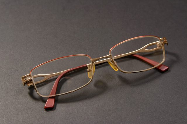 Eyeglasses rest on a dark surface, featuring golden frames with red temple tips and clear rectangular lenses, positioned unfurled and inactive.
