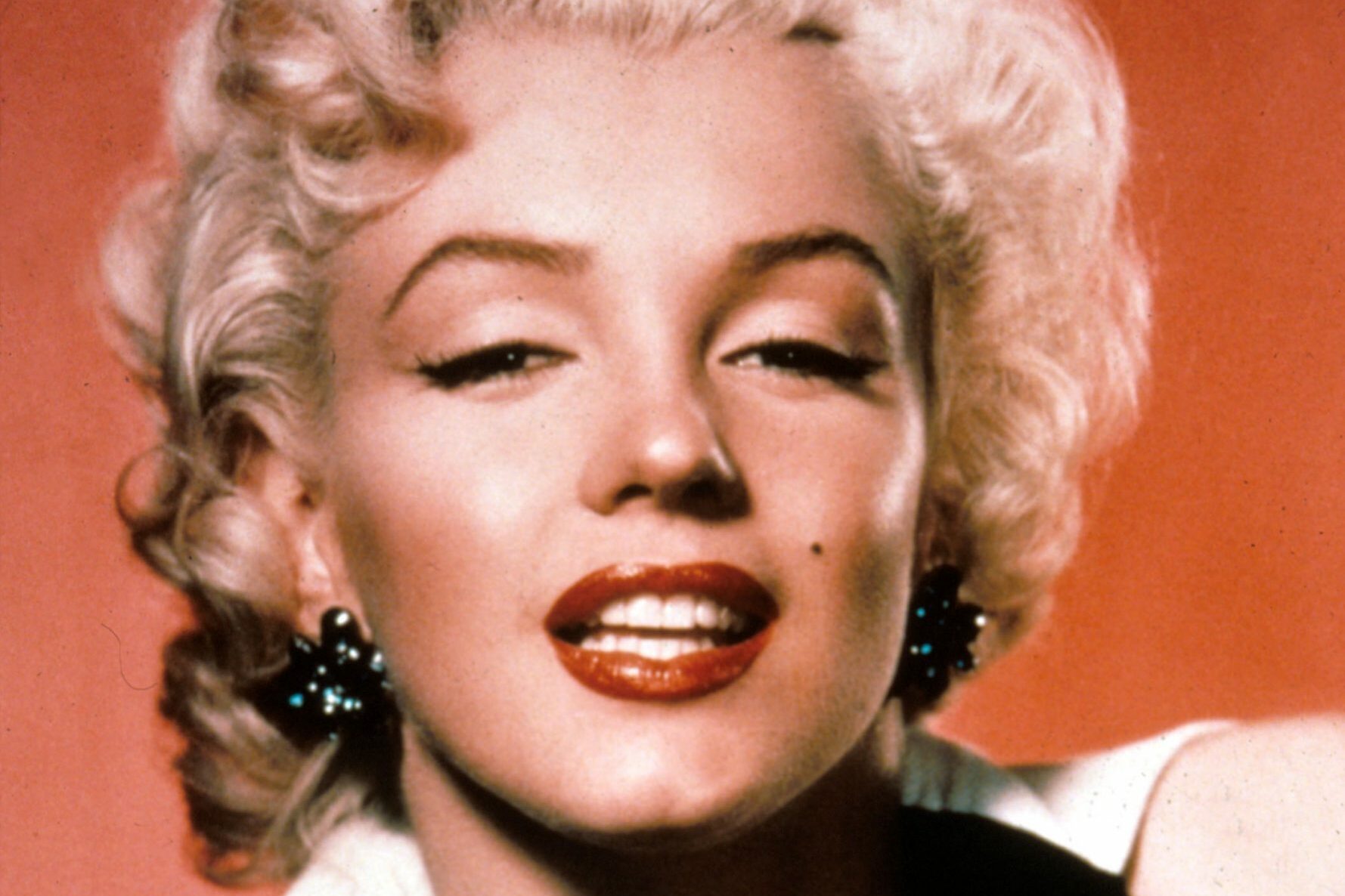 A woman with blonde, curly hair smiles slightly, wearing bright red lipstick and black earrings, against a warm orange background.