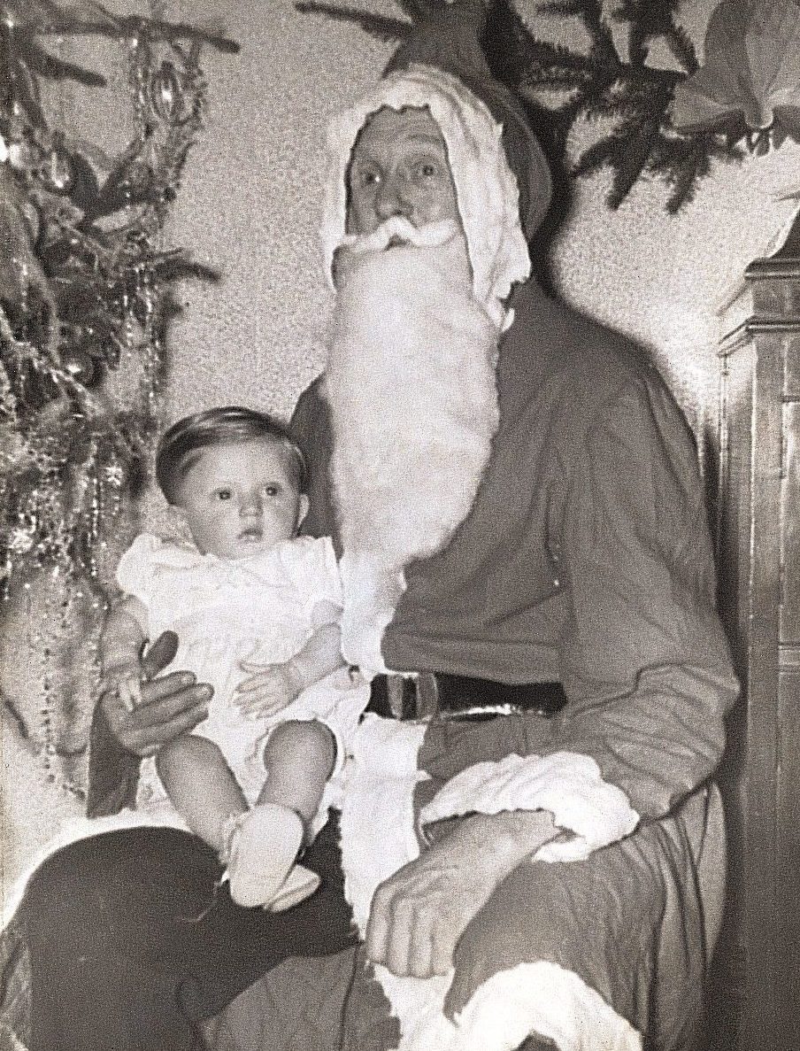Santa holds a small child on his lap, surrounded by Christmas decorations in a festive setting.