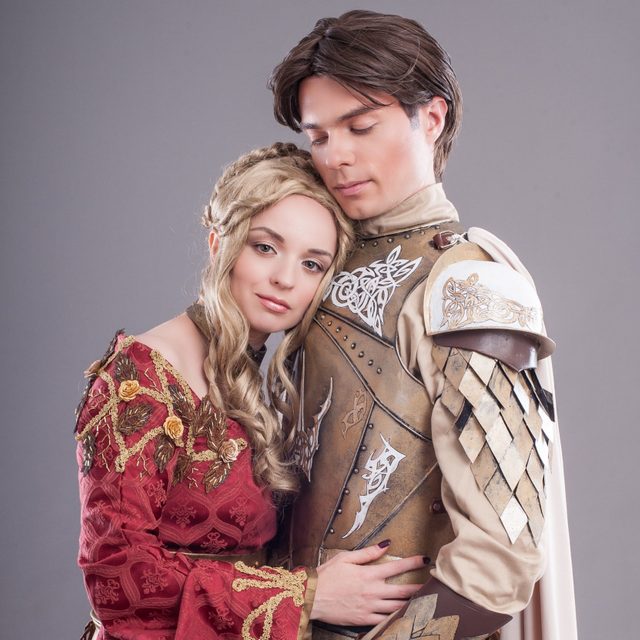 A woman in an ornate red dress embraces an armored man, both appearing serene, set against a neutral gray background.