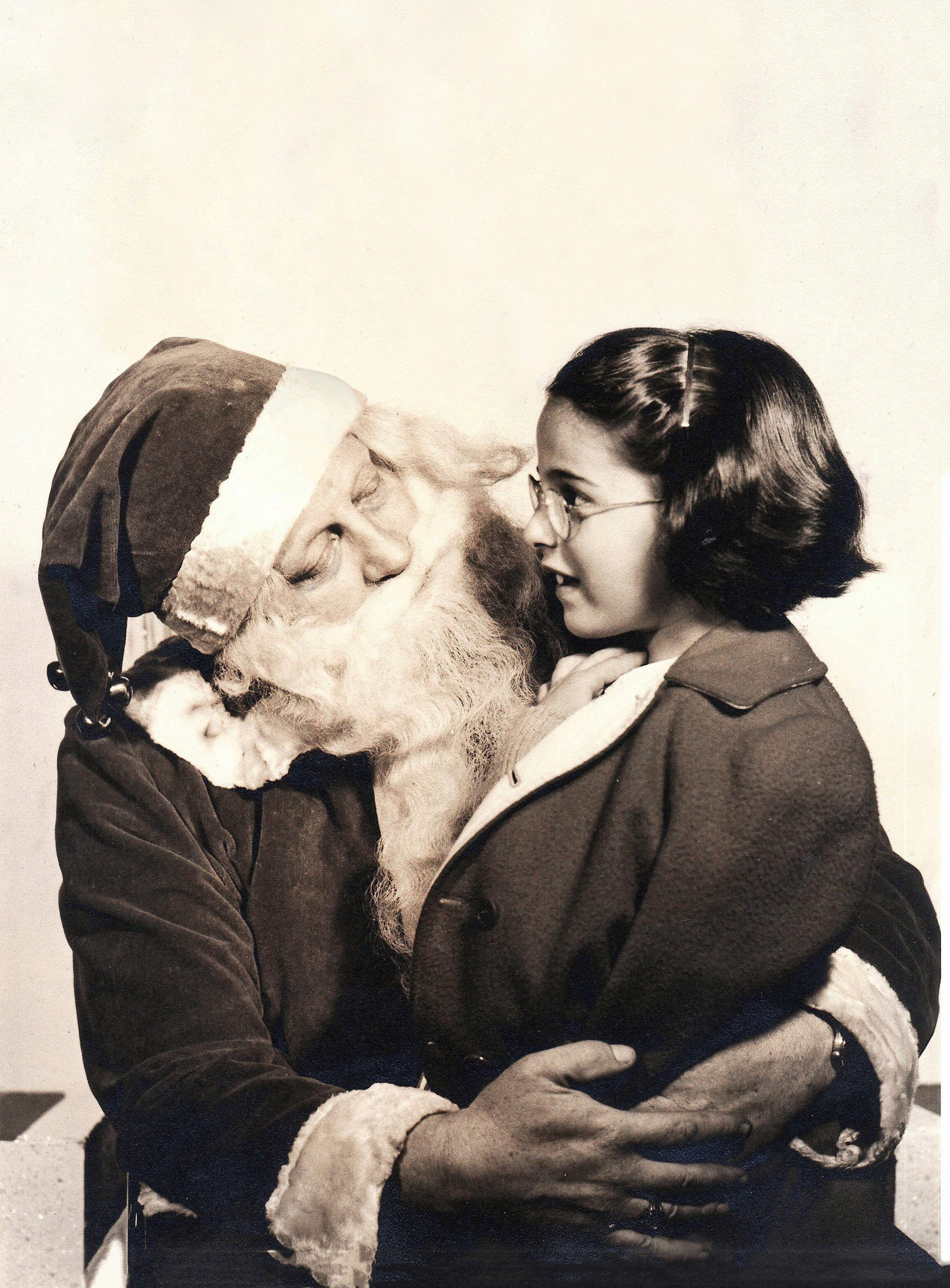 Santa Claus, smiling, holds a young girl in his arms. They are exchanging a joyful glance in a festive setting.