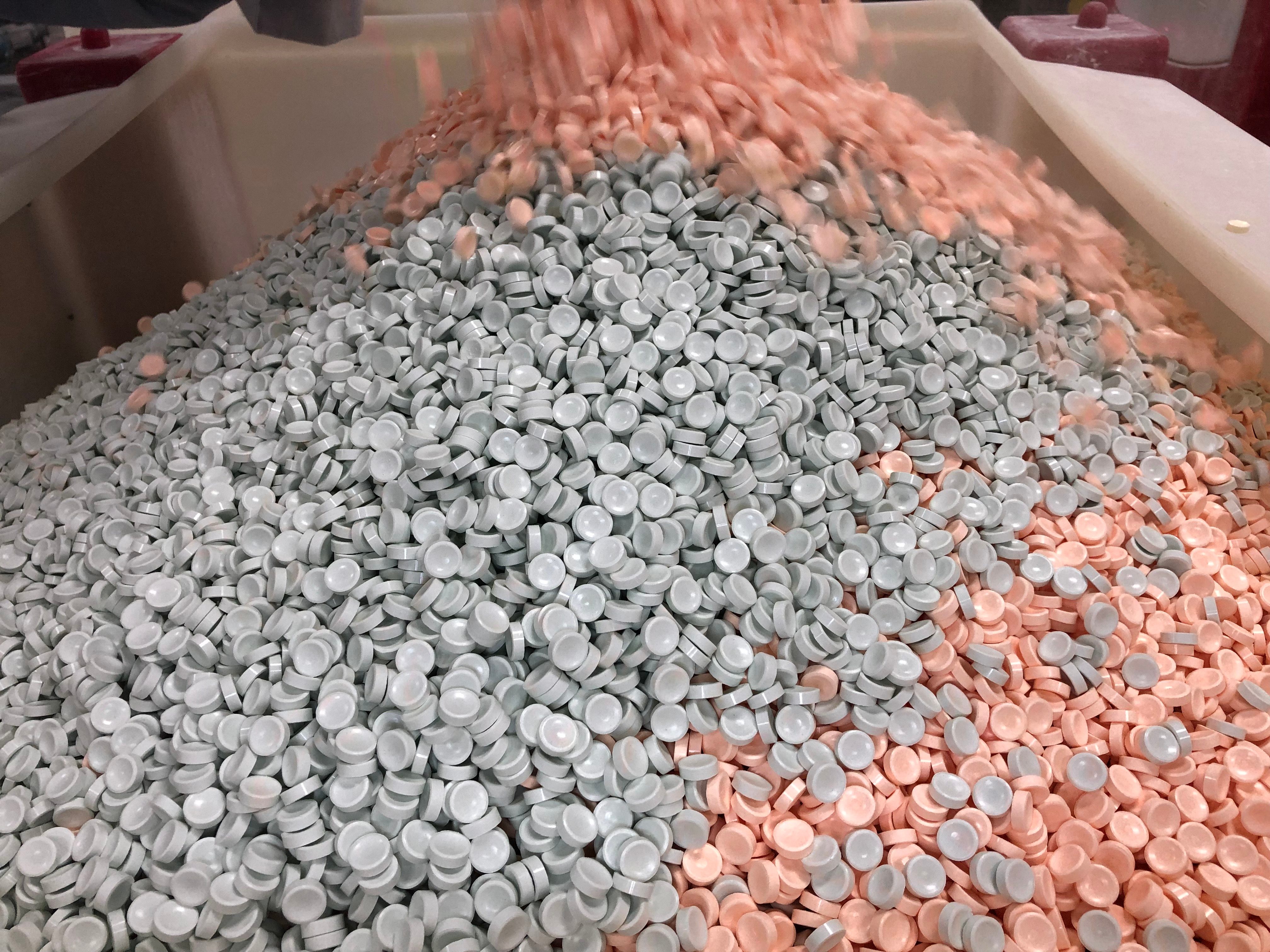 Many small, round plastic caps cascade into a large tray, mixing gray and peach colors, amid industrial machinery.