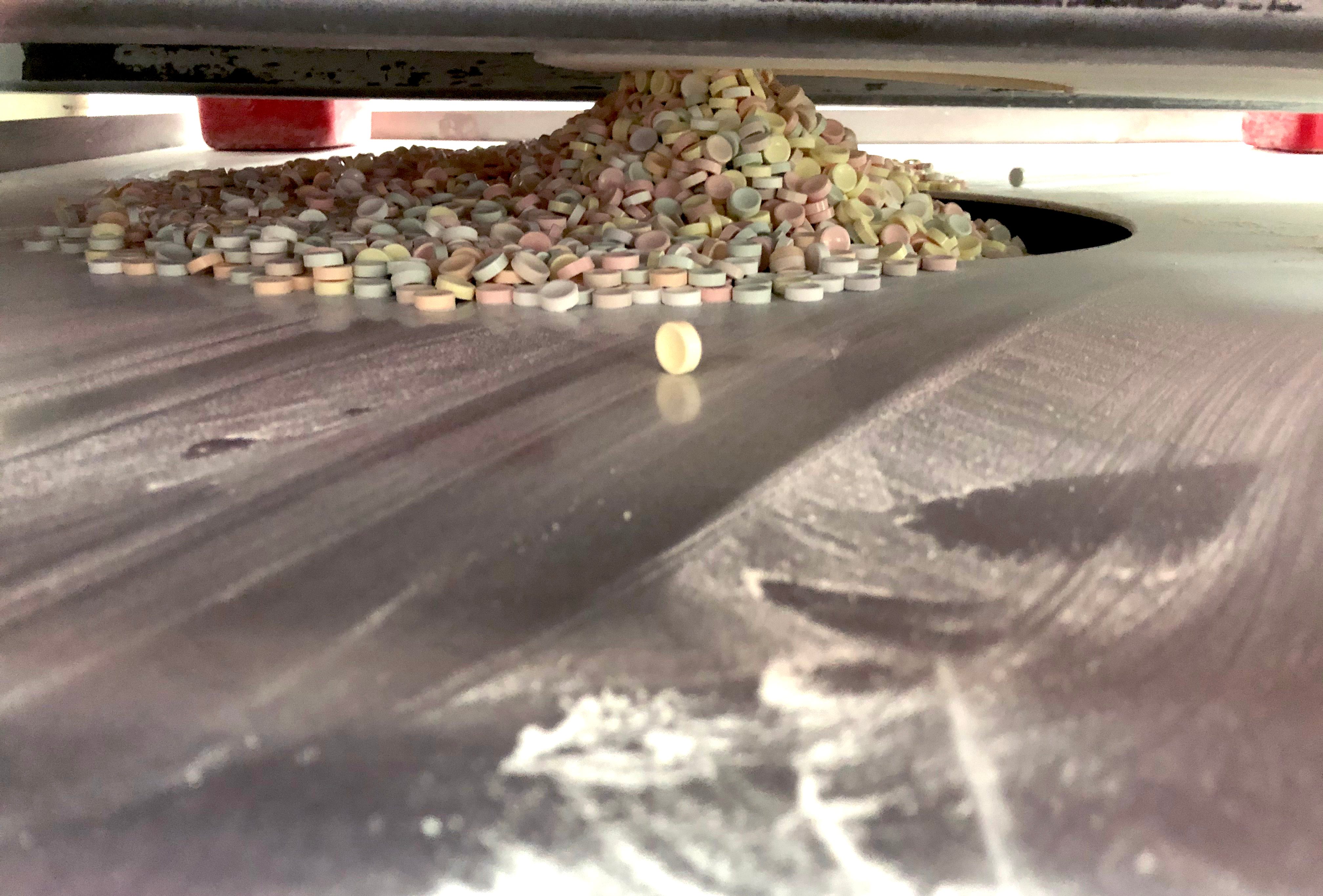 Pills spill onto a dusty, metallic surface, forming a pile under a machine in an industrial setting.