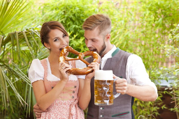 Pretzel being shared by smiling couple, man holding beer stein; lush green foliage in background.