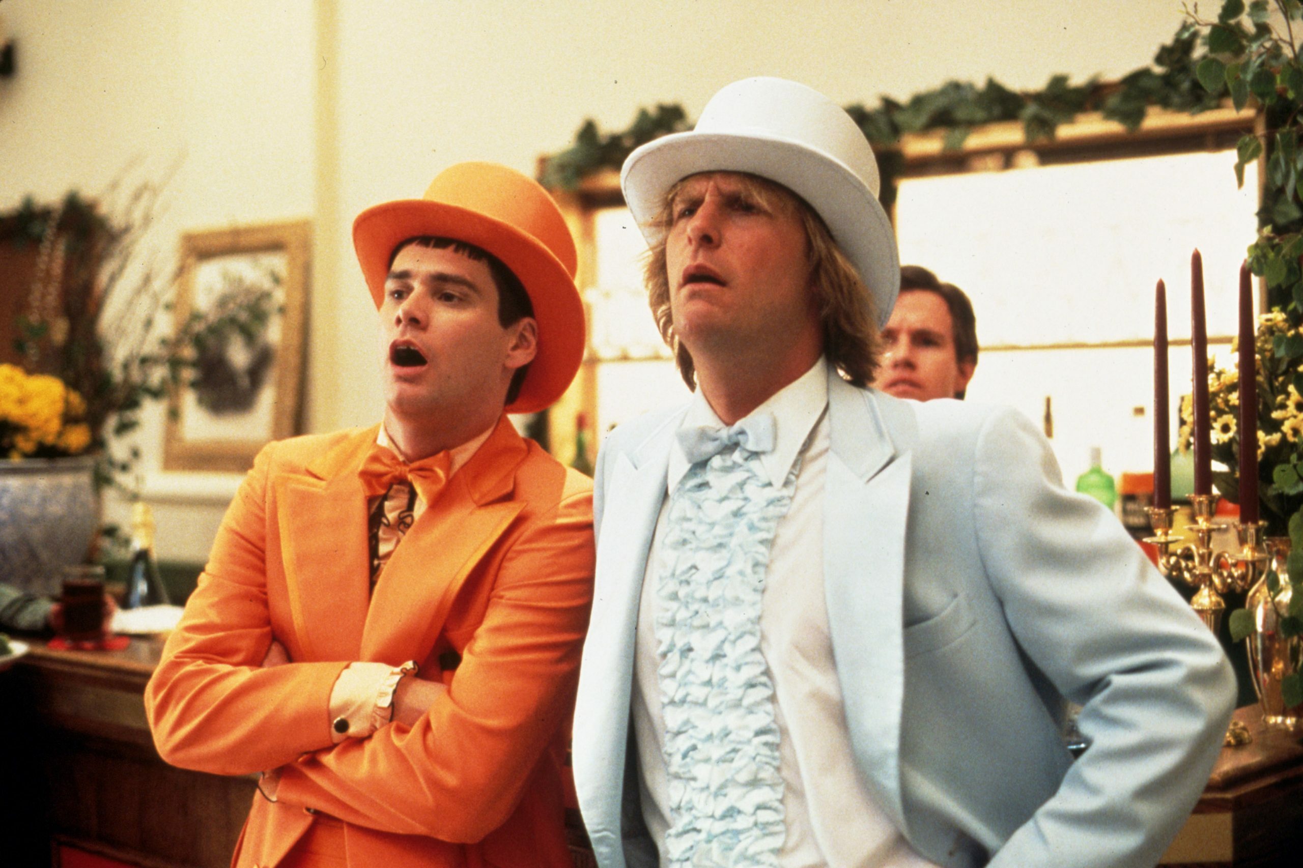 Two men wearing orange and blue tuxedos stand surprised at a fancy indoor event with floral decorations and a bar in the background.