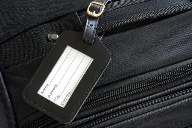 A luggage tag hangs on a black suitcase, featuring spaces for Name, Address, City, and Country.