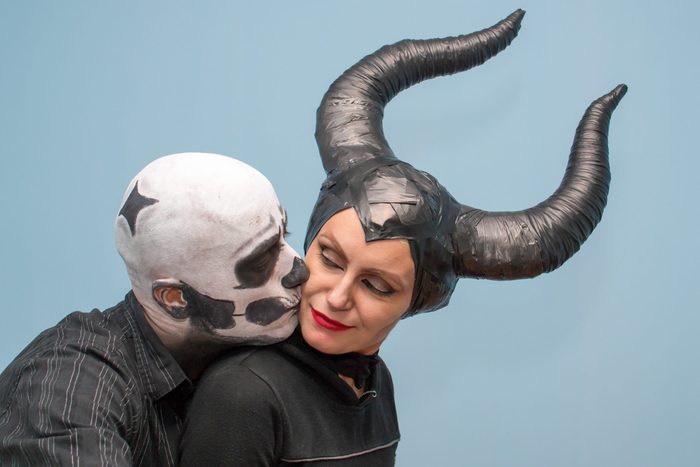 Person in skull makeup leans towards another wearing black horns, both on a plain blue background.