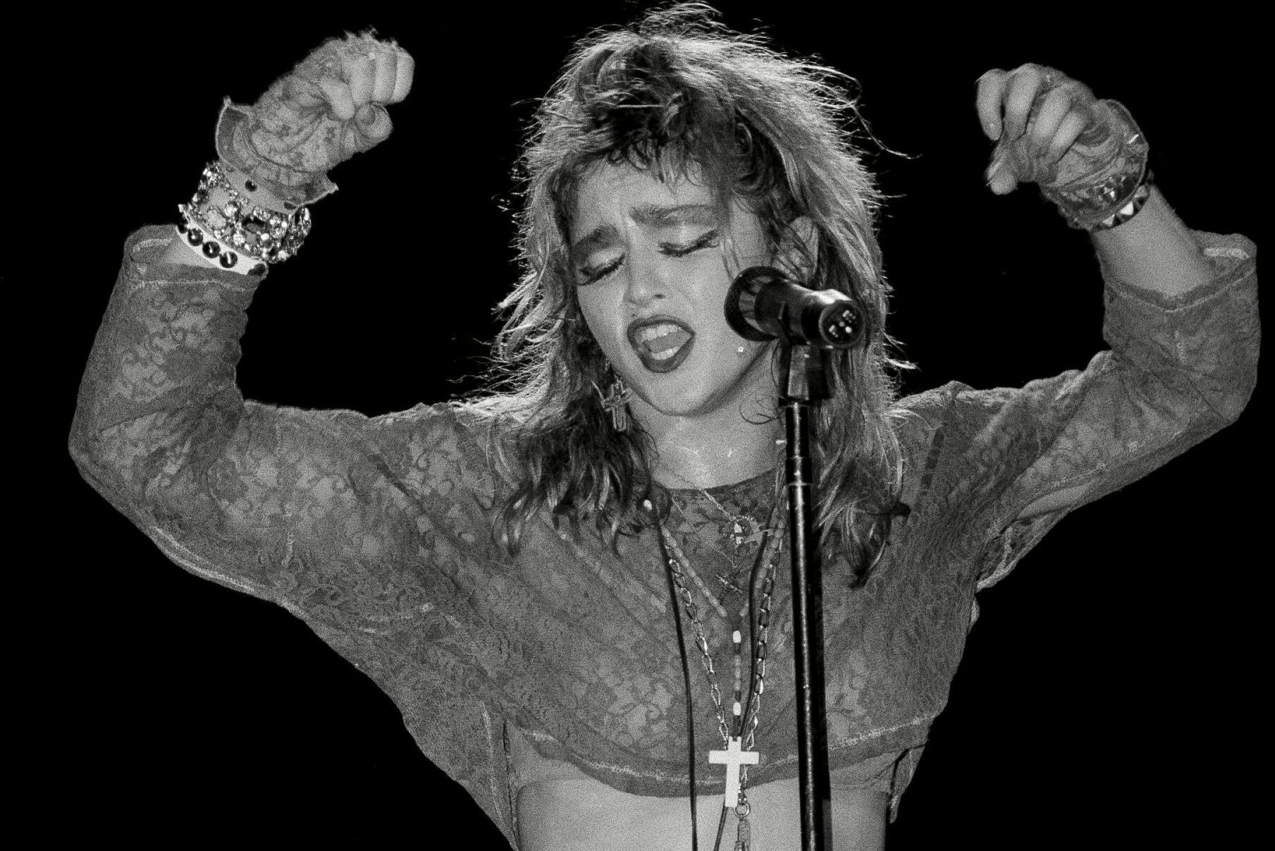 Singer passionately performs on stage, raising fists while singing into a microphone, wearing layered jewelry and a lacy outfit against a dark backdrop.