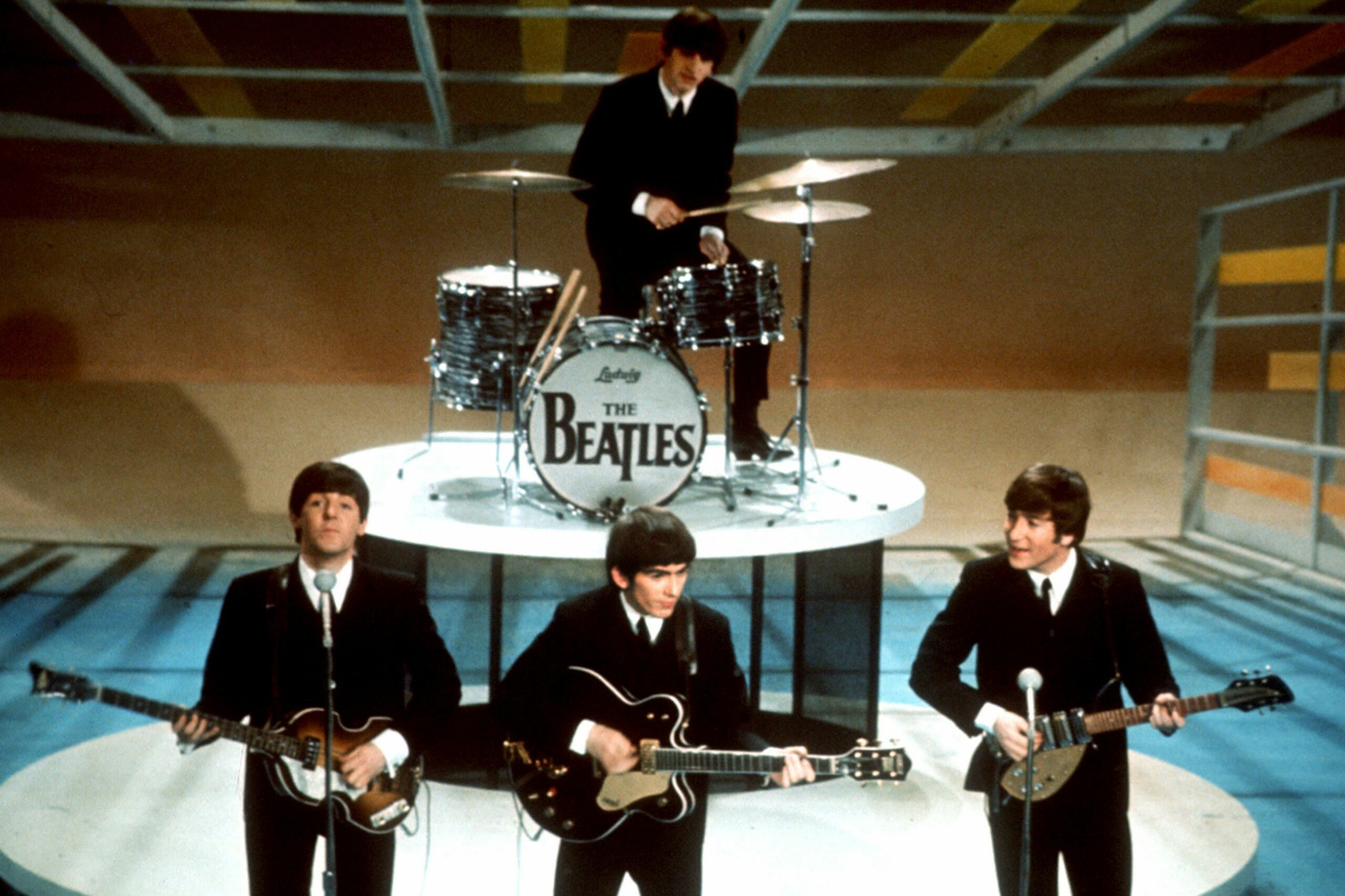 Three musicians play guitars on stage; behind them, a drummer sits with a drum labeled "Ludwig The Beatles." The stage has colorful geometric patterns.