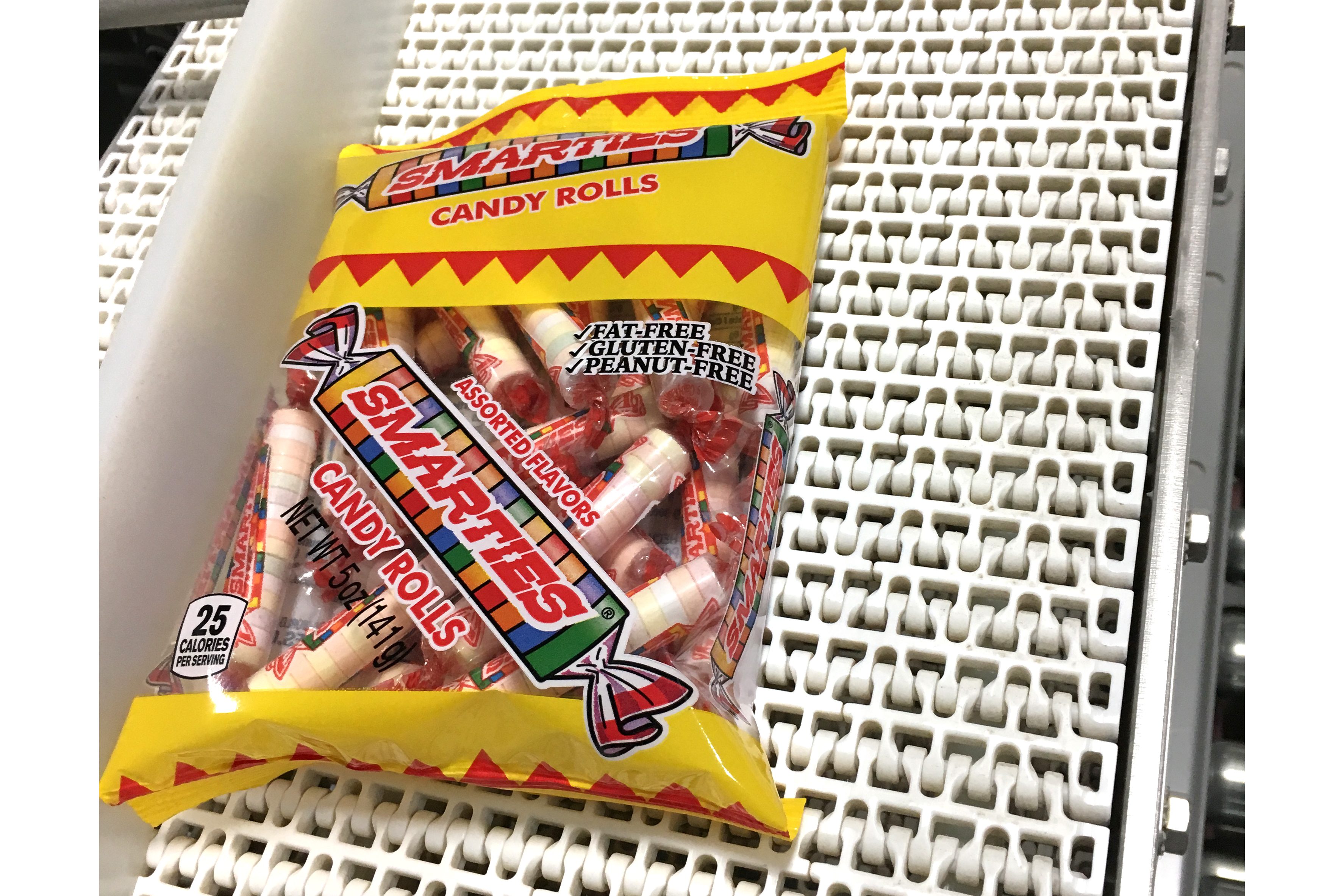 Bag of Smarties candy rolls sits on a white conveyor belt, displaying labels for fat-free, gluten-free, peanut-free candy.