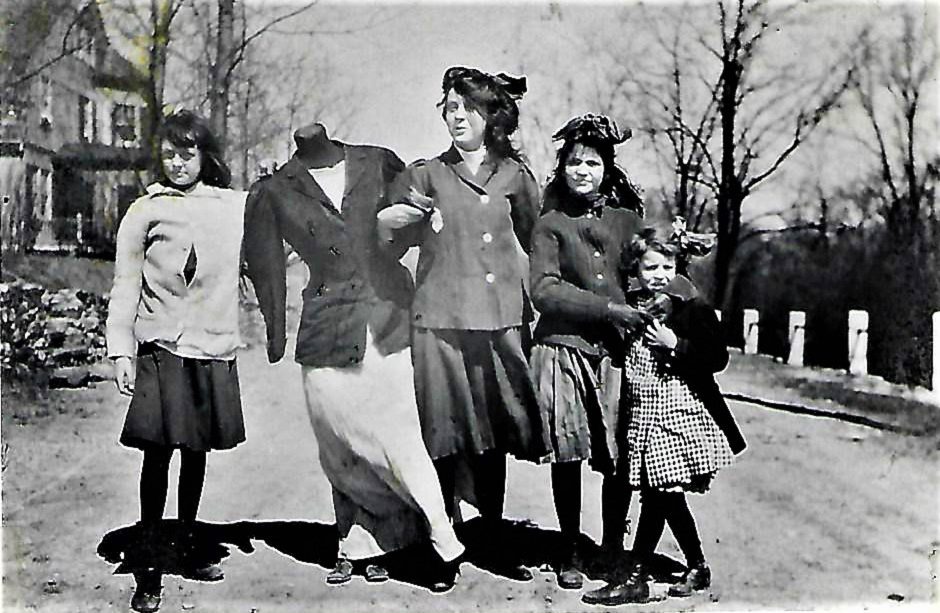 Five girls pose on a rural road, linked arm in arm, with one headless figure among them, barren trees in the background.