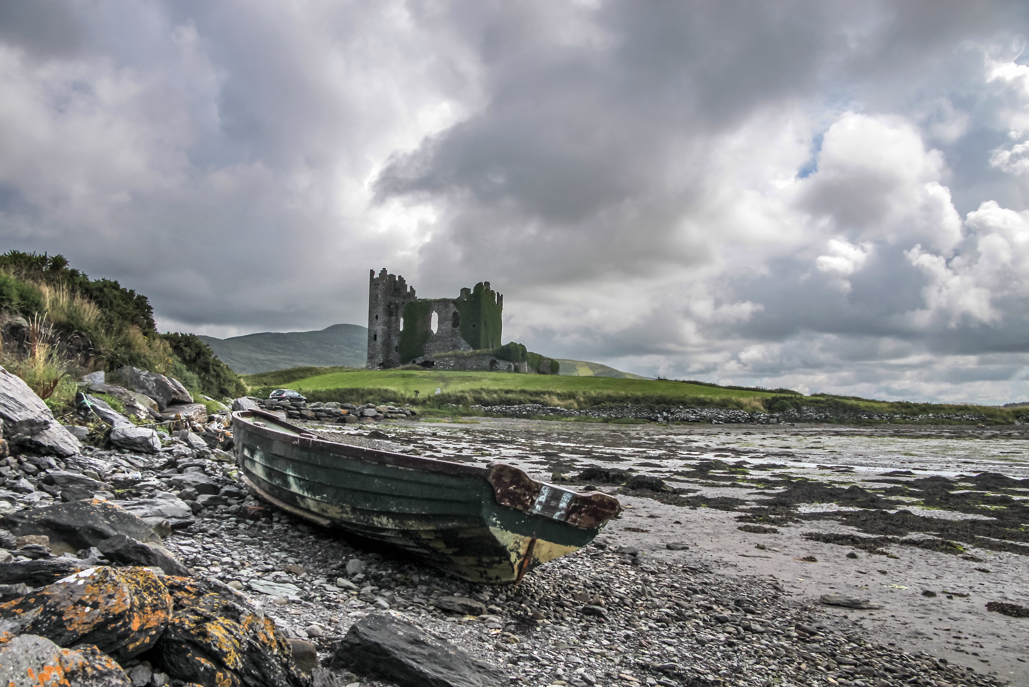 A weathered boat rests on rocky shore, with an ancient stone castle in the background under a cloud-filled sky.