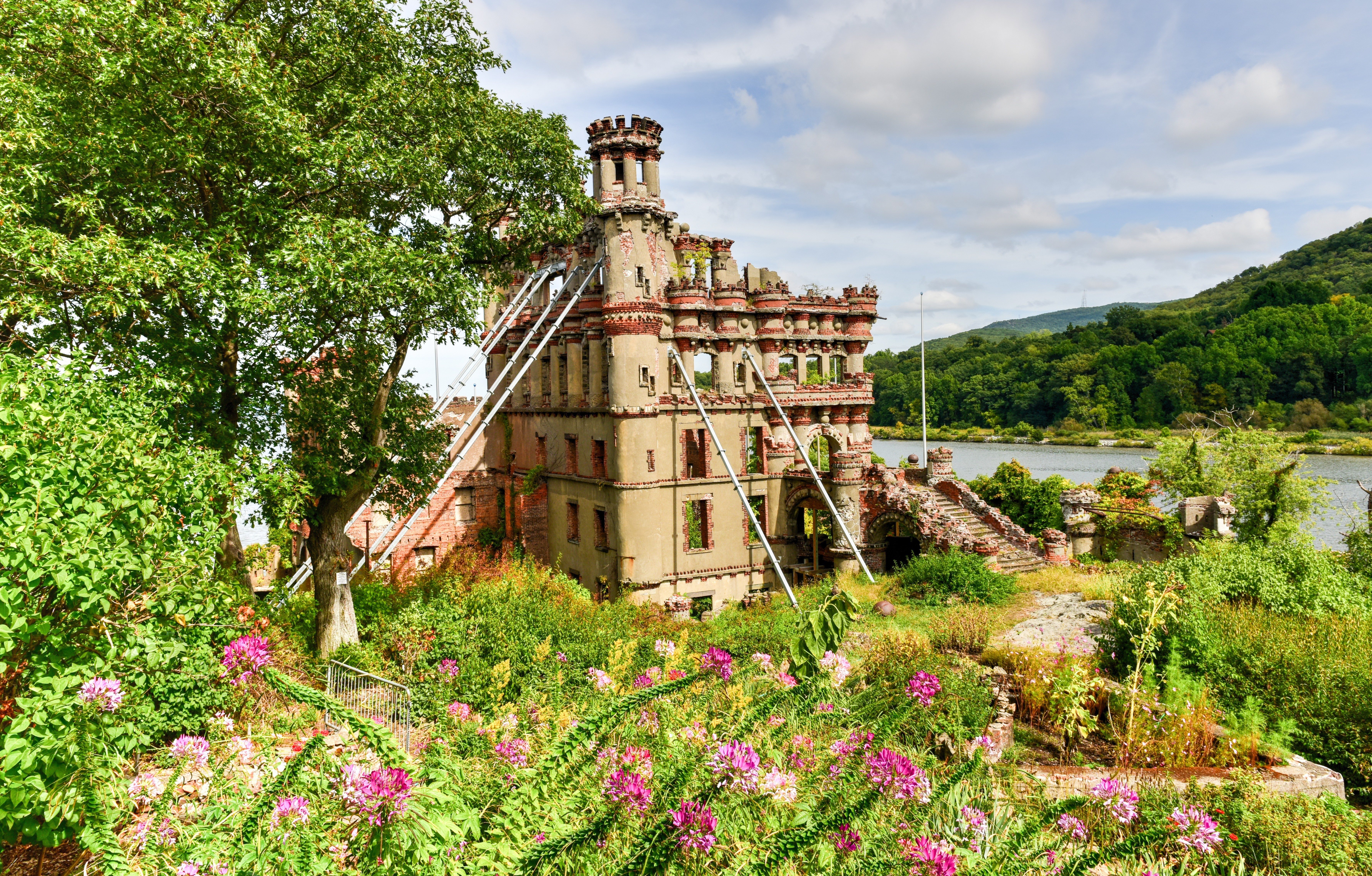 Ruined castle sits amidst vibrant flowers; scaffolding supports the structure. A river flows nearby, bordered by lush green hills under a cloudy sky.