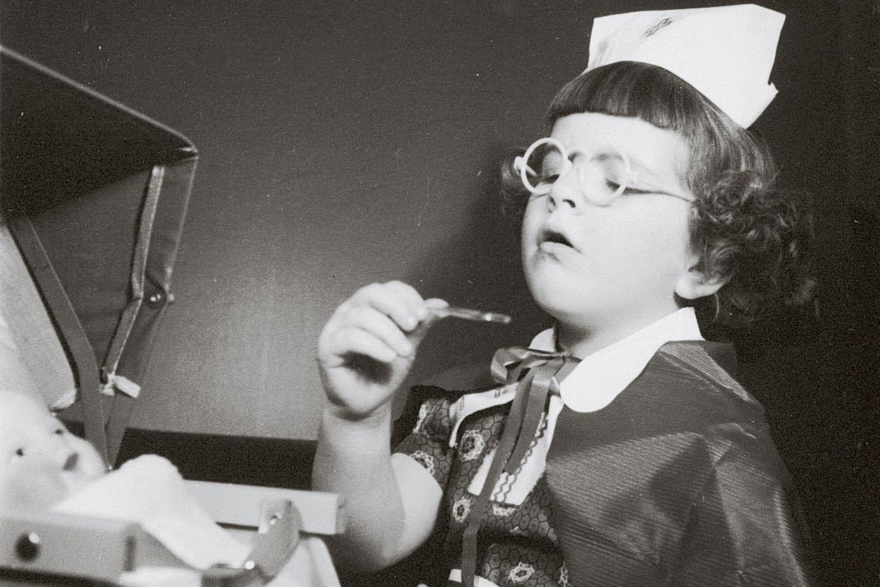 Child wearing glasses and a nurse hat examines a toy doll with a thermometer, set in a play medical scene.