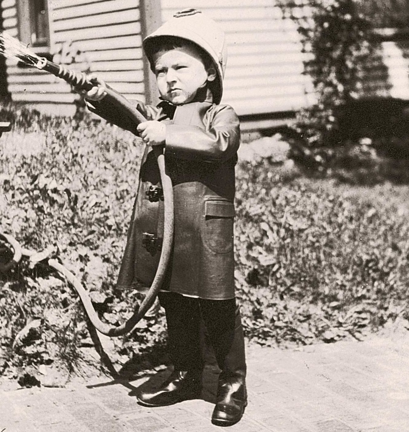 Child sprays water with a hose, wearing a firefighter outfit, standing on a garden path near a house surrounded by plants.