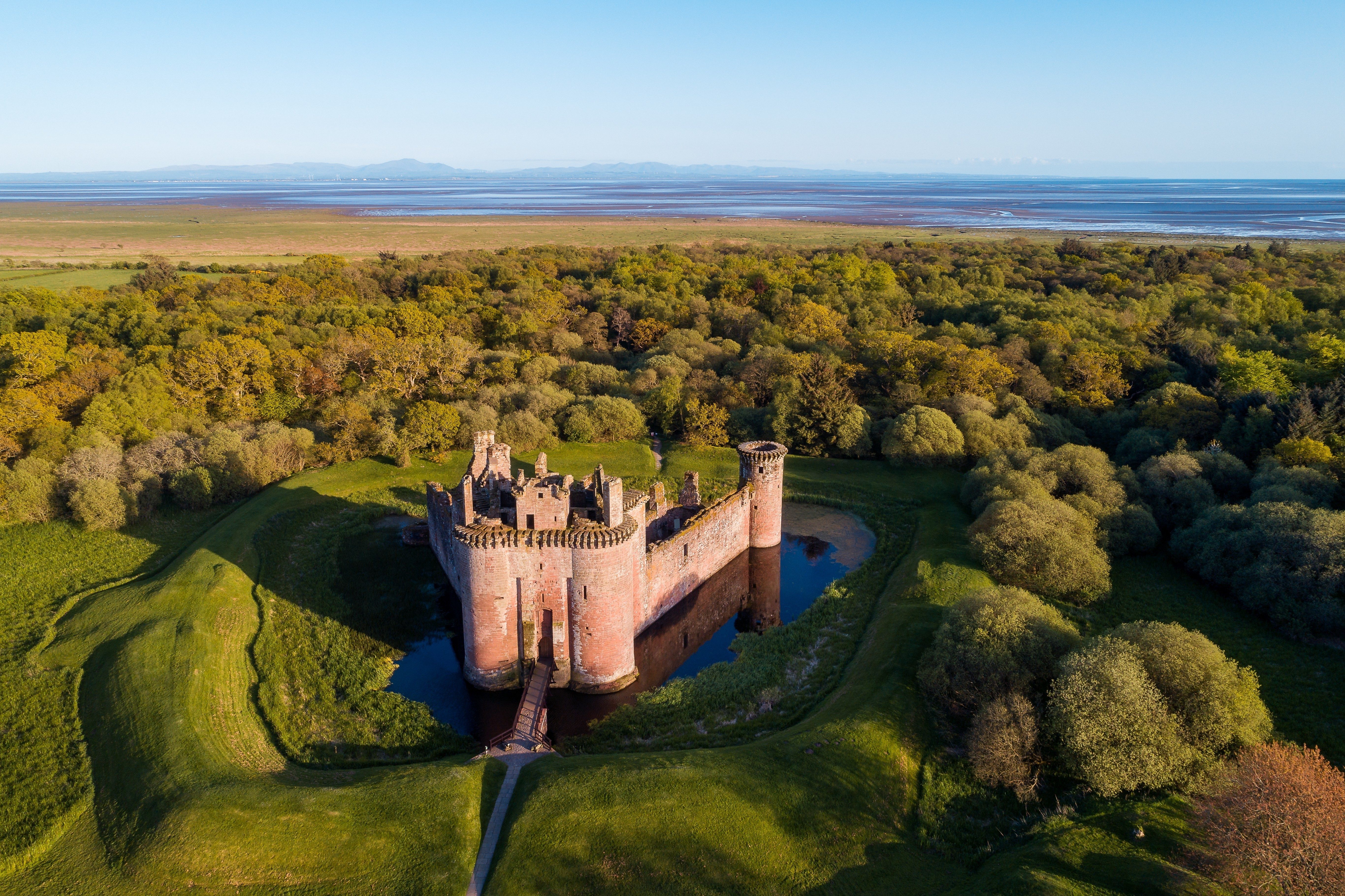 A stone castle surrounded by a moat, situated amidst lush greenery and dense trees with a distant view of the coastline.