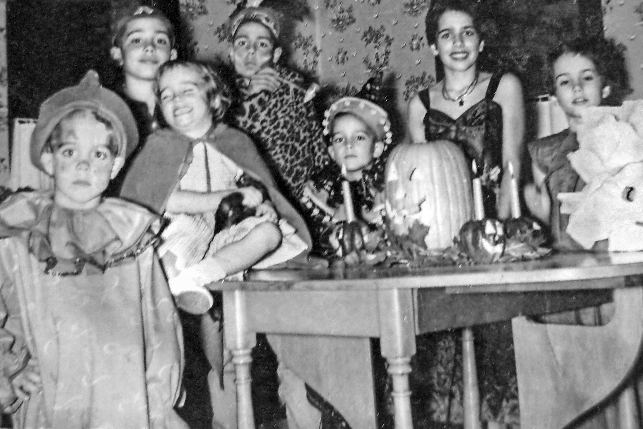 Children in costumes pose around a table with carved pumpkins and candles, in a festive setting.