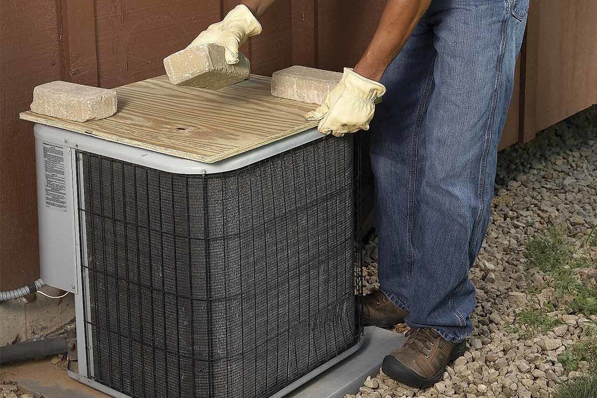 Air conditioning unit topped with a board, held down by bricks, surrounded by gravel near a wooden structure. Person positioning a brick.