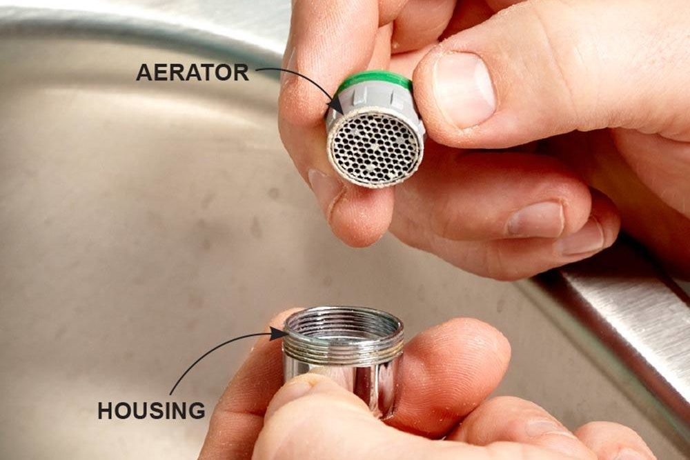 Hands holding a faucet aerator above its housing, removing it for cleaning, with a kitchen sink in the background. Labels: "Aerator" and "Housing".