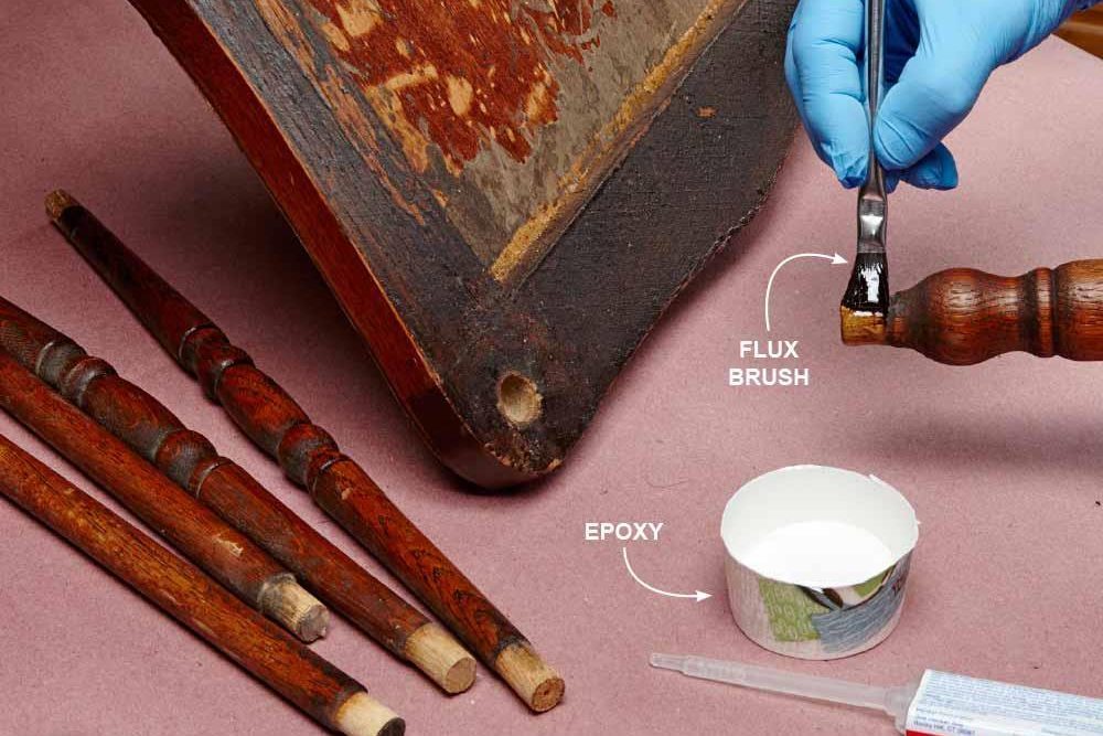 Brush applying epoxy to wooden chair leg, surrounded by dismantled spindles on a tabletop. Text labels indicate "FLUX BRUSH" and "EPOXY."