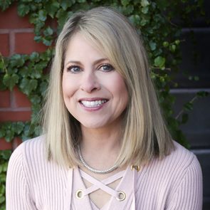 A woman smiles outdoors, wearing a pink sweater and necklace, with a green ivy-covered brick wall in the background.