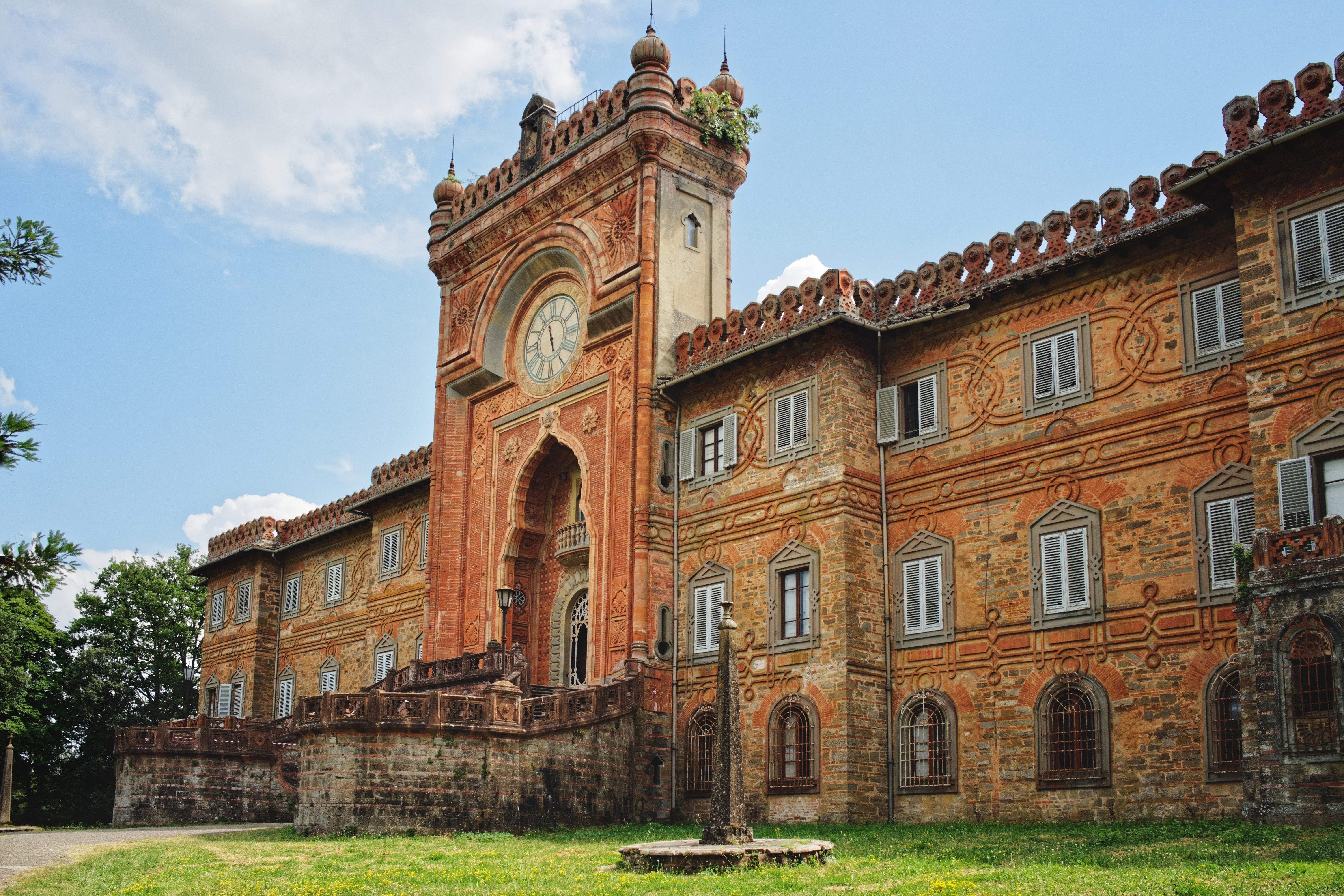 Ornate brick building features a clock tower and intricate arches, set in a grassy area with trees and clear blue sky background.