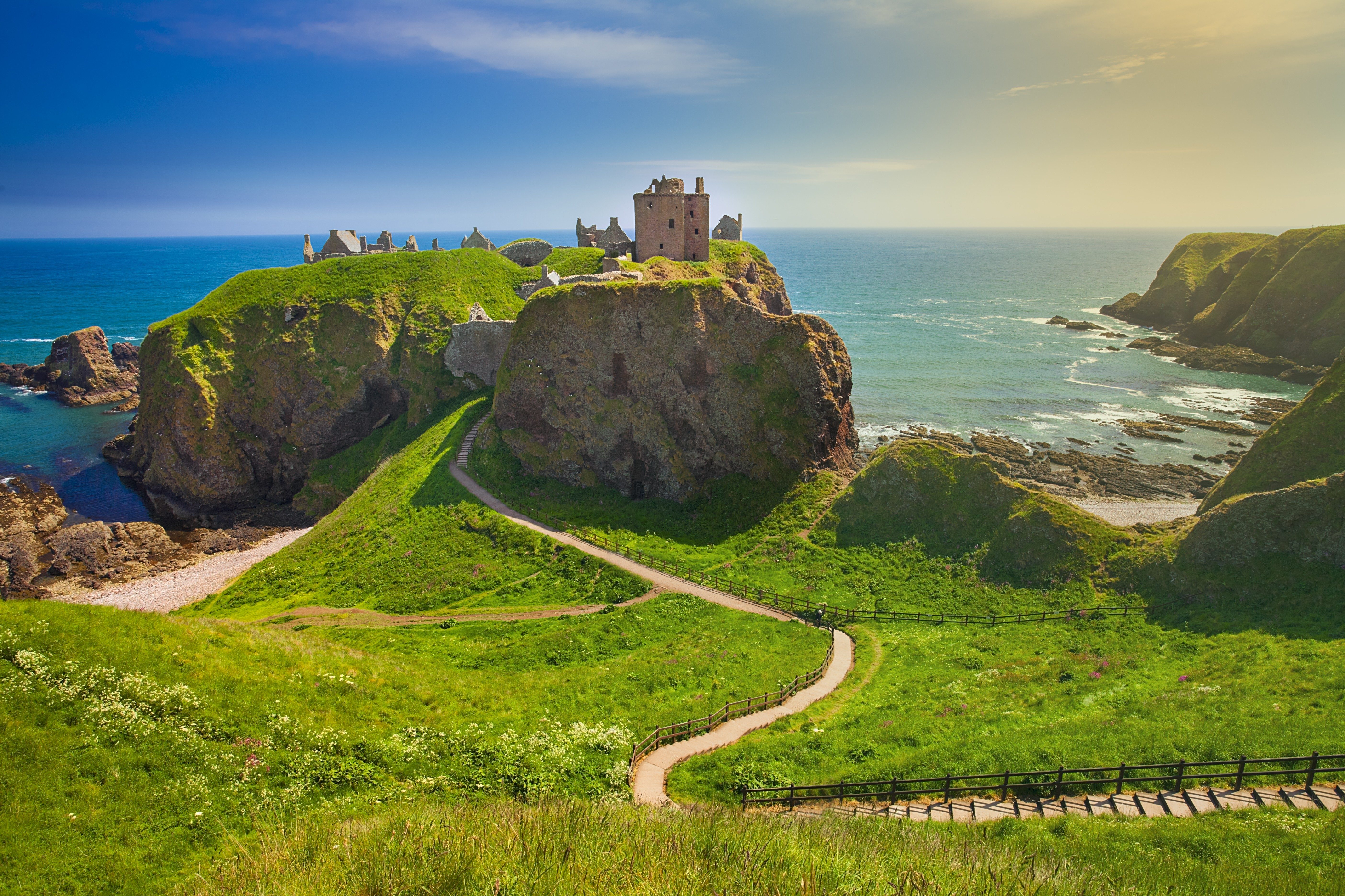 Castle sits on a grassy cliff, overlooking the sea, connected by a winding path amidst lush hills under a clear sky.