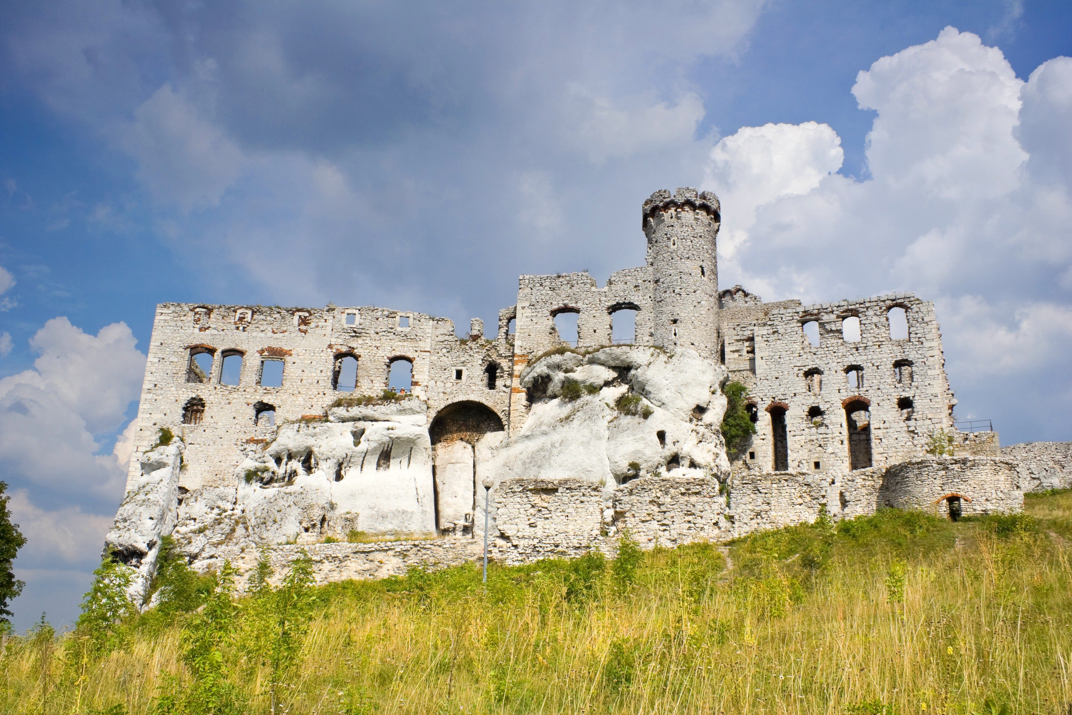 Stone castle ruins stand on rocky hill under a partly cloudy sky, surrounded by grass and brush.