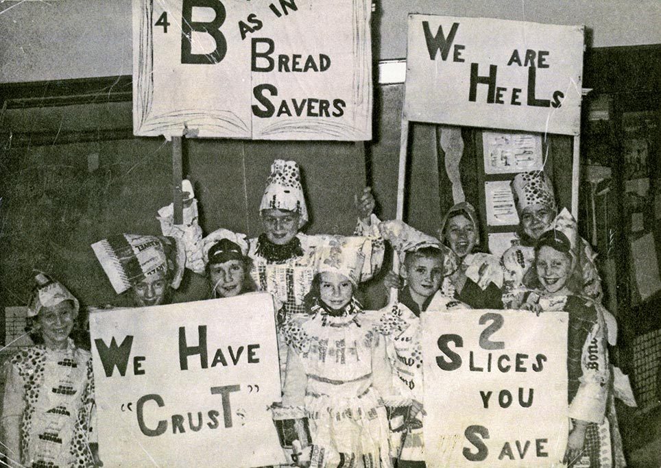 Children dressed in hats and outfits hold signs with bread-related puns, smiling in a school setting.