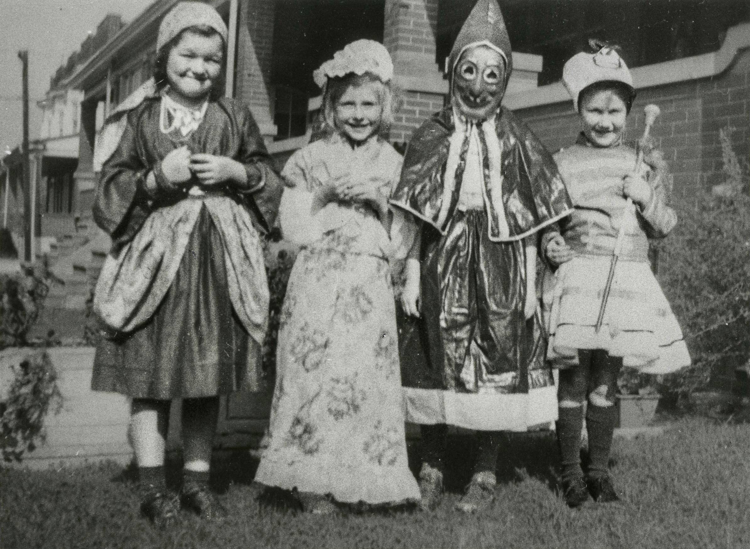 Four children wear vintage costumes, standing in a row, outdoor setting with houses visible in the background.