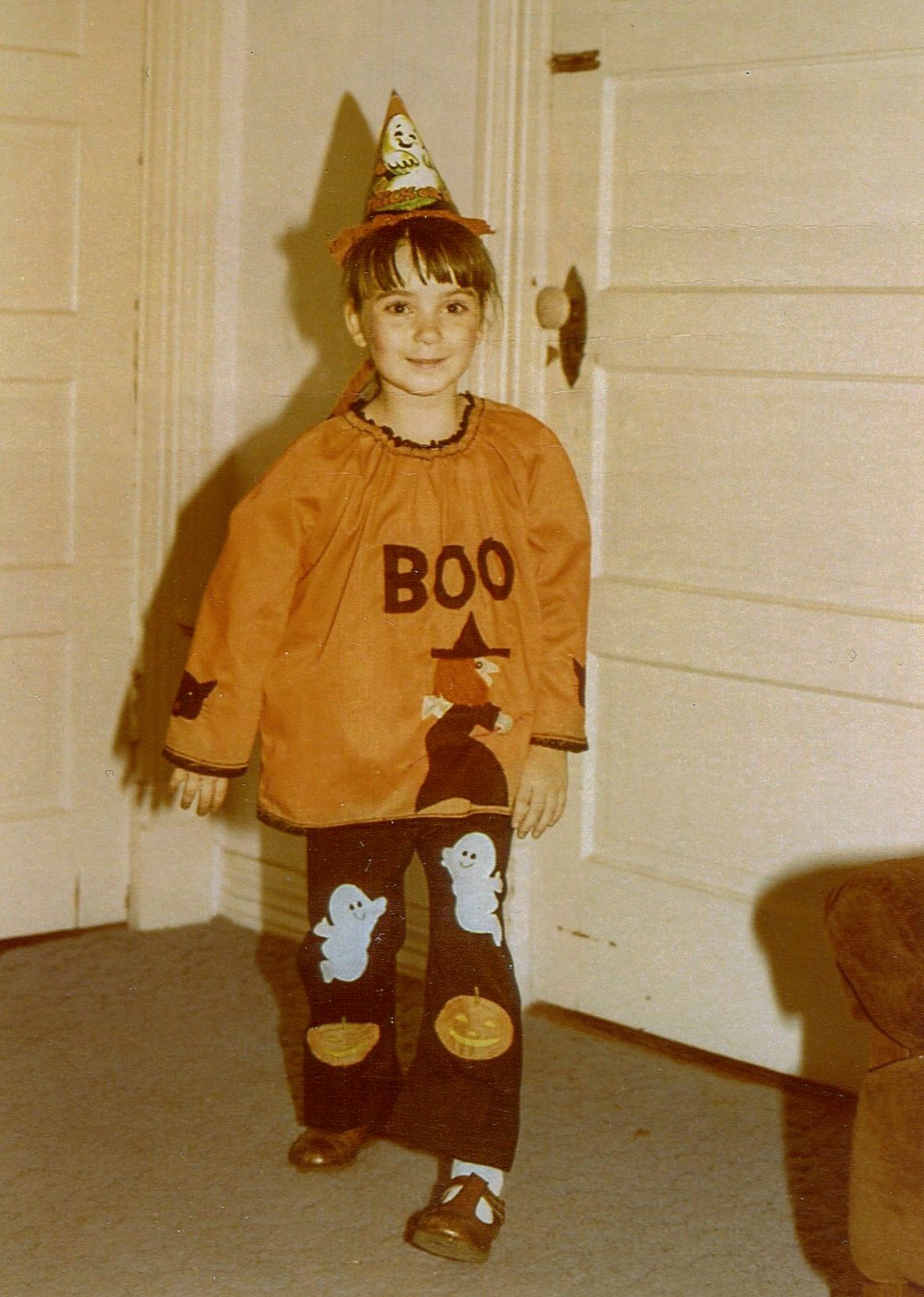 Child wears orange costume reading "BOO," decorated with witch, ghosts, and pumpkins, inside a room with white doors and a couch.