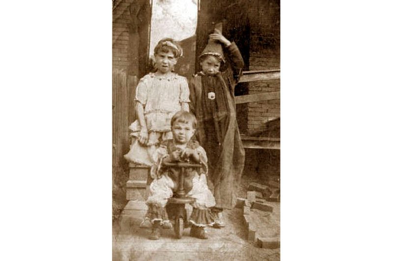 Three children pose outdoors; one sits on a tricycle, while the others stand behind, near a house entrance surrounded by bricks.