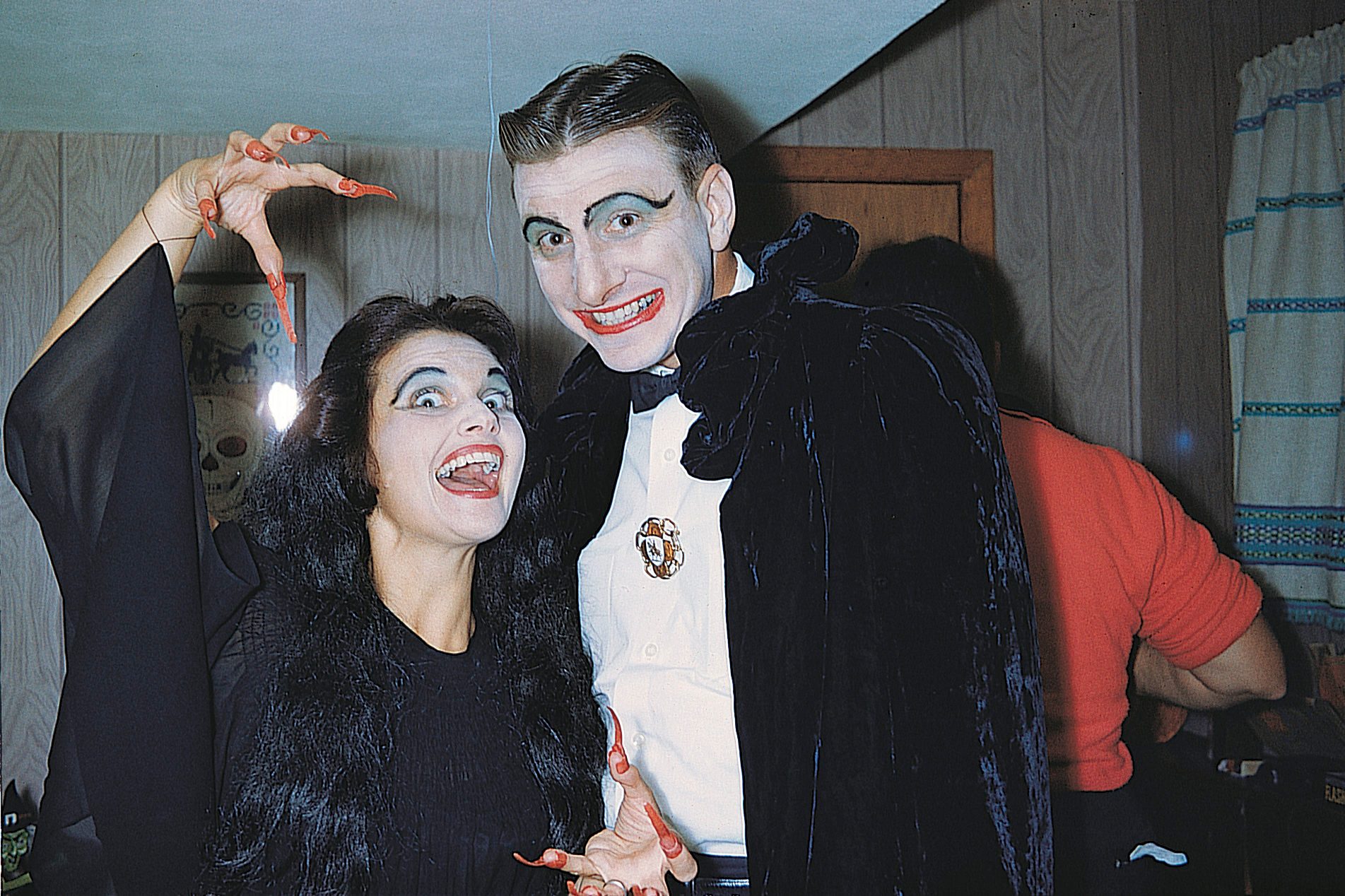 Two people in dramatic makeup pose playfully in a room with wood-paneled walls, one gesturing with claw-like hands and smiling widely.