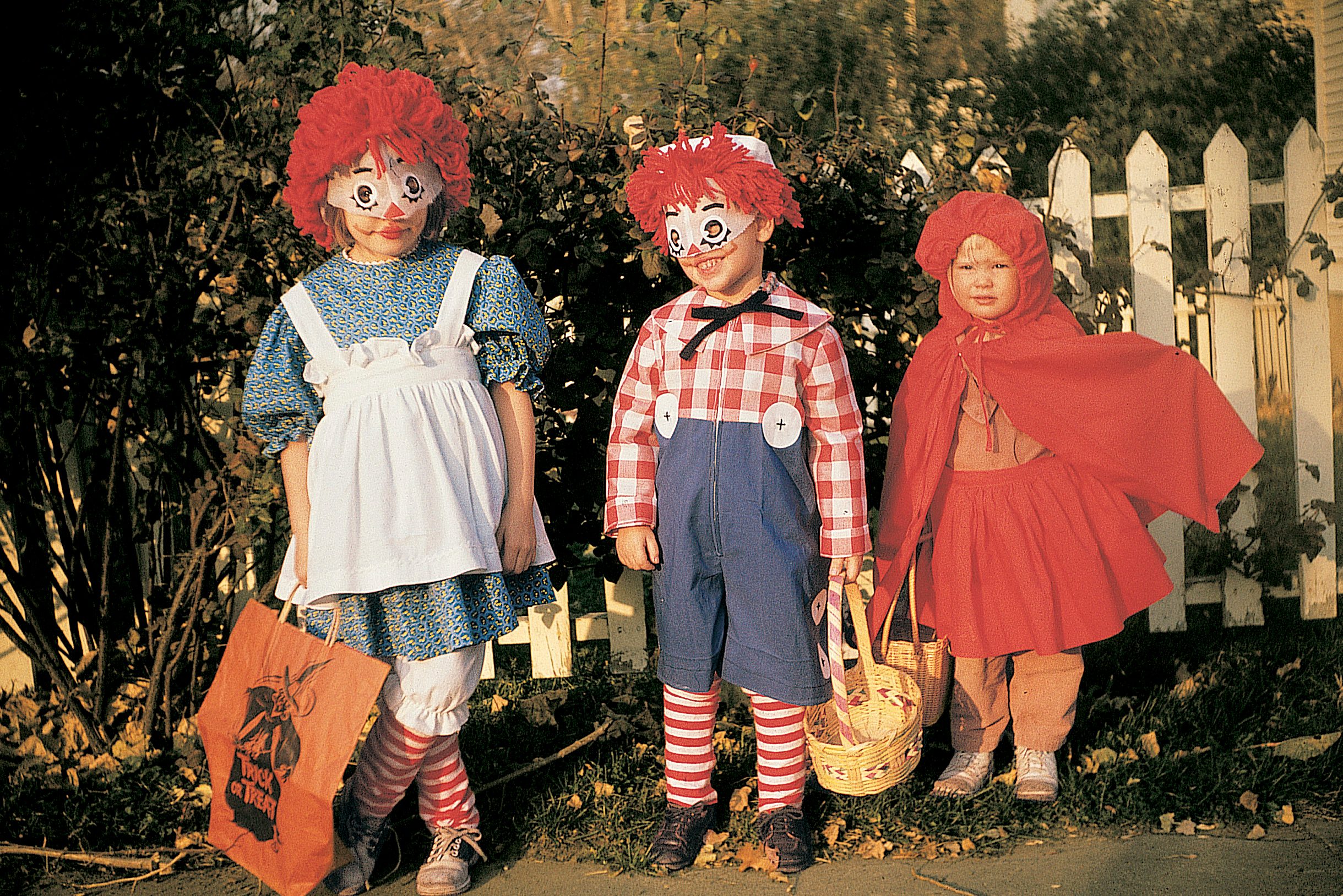 Children stand in Halloween costumes holding bags, in front of a white picket fence and bushes.