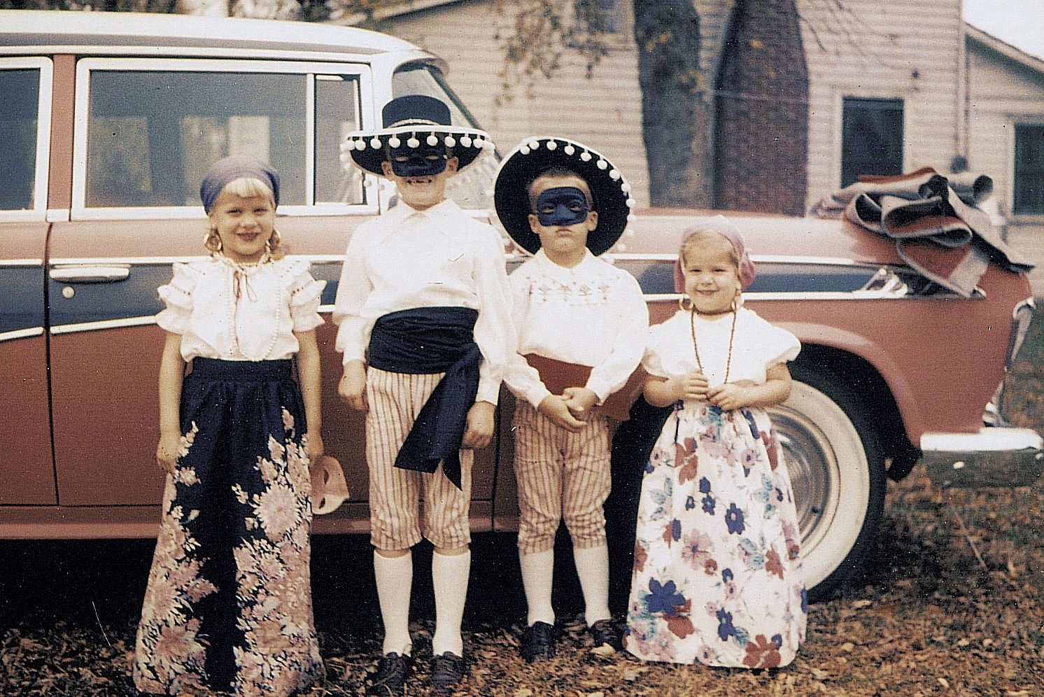 Four children stand wearing festive costumes, featuring hats and masks, in front of a vintage car parked outside a house.