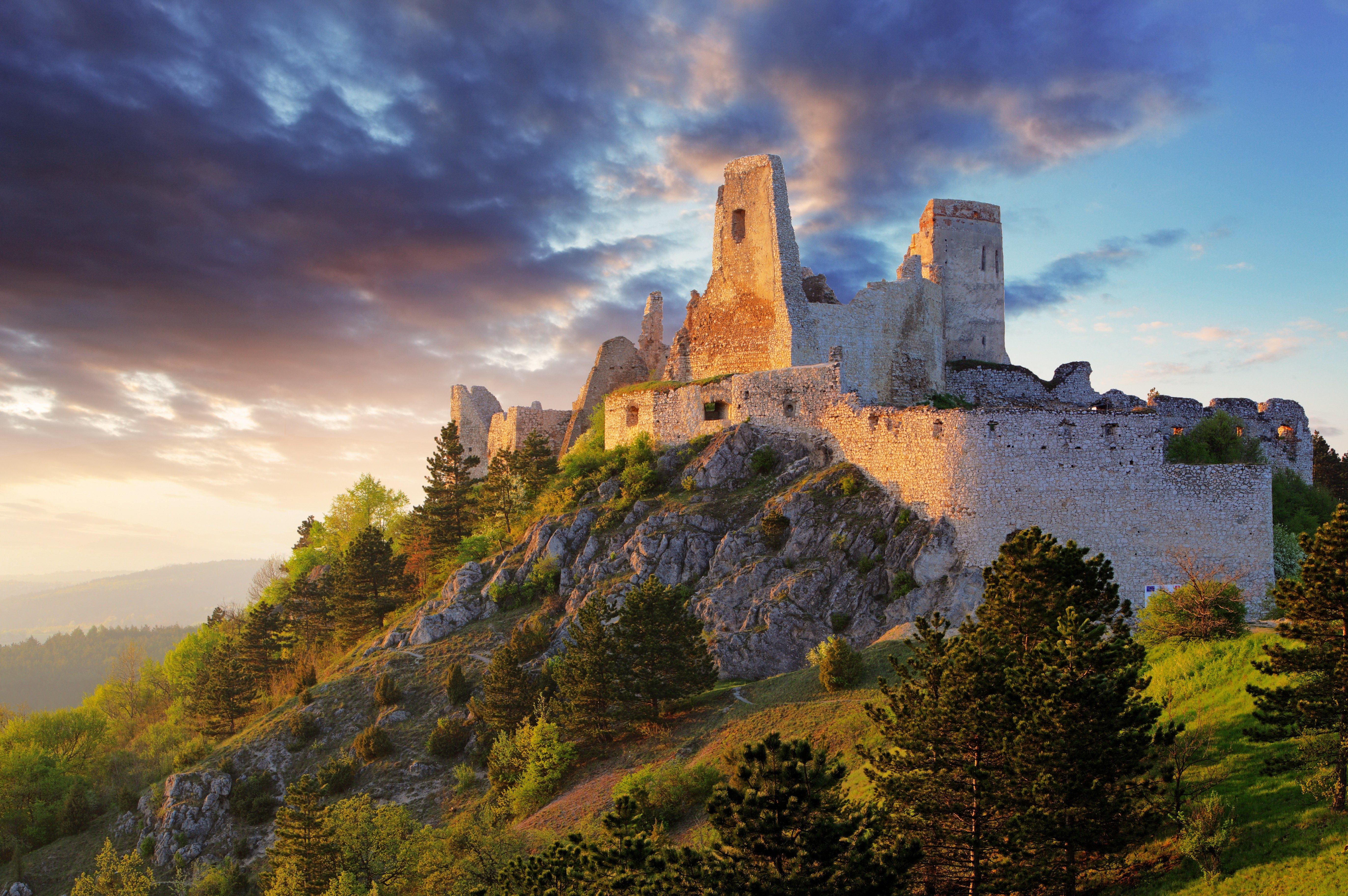 Ancient castle ruins stand atop a rocky hill, surrounded by dense green trees, under a dramatic, colorful sky at sunset.