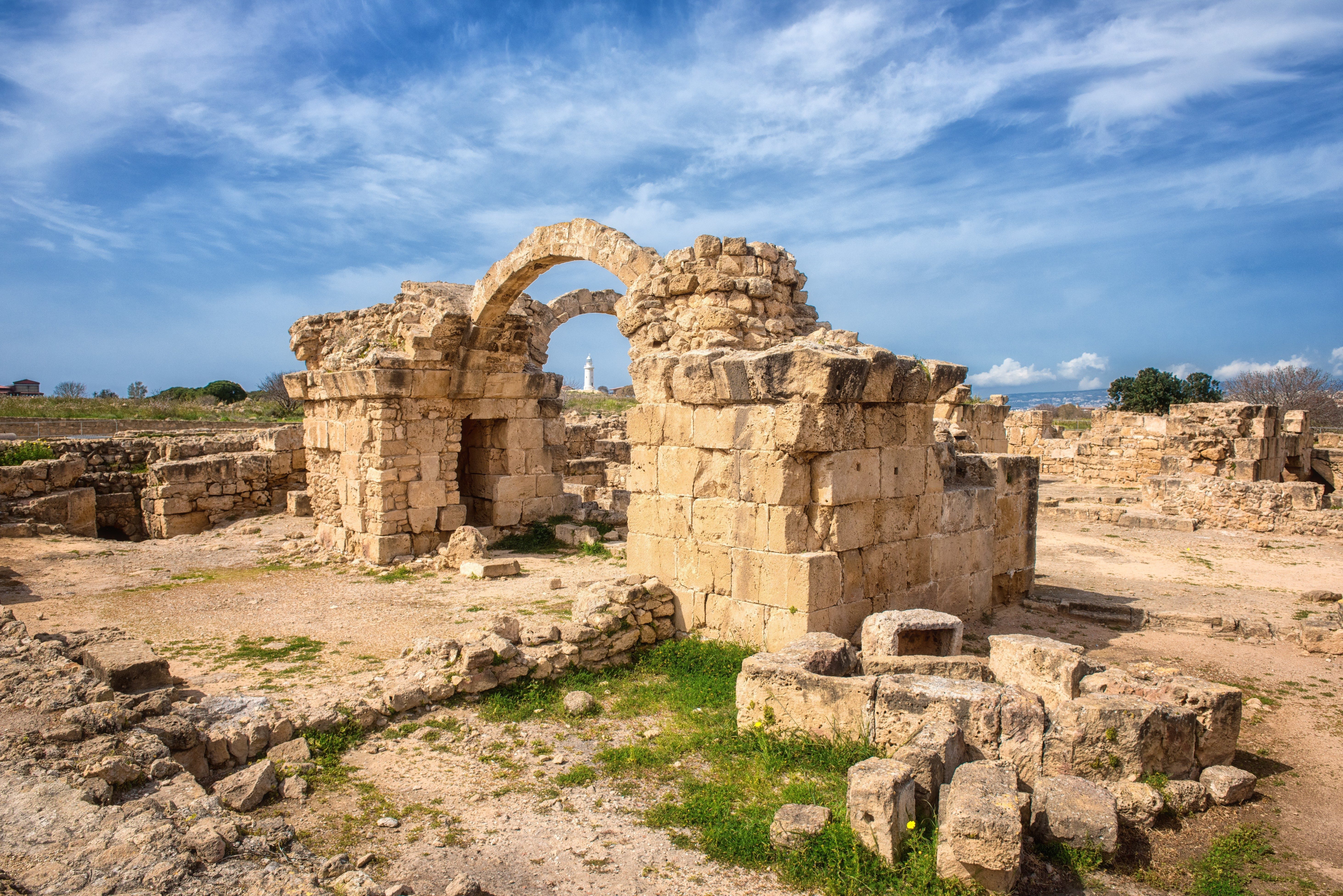 Ancient stone ruins stand amidst grassy terrain under a vibrant blue sky, with a distant lighthouse visible on the horizon.