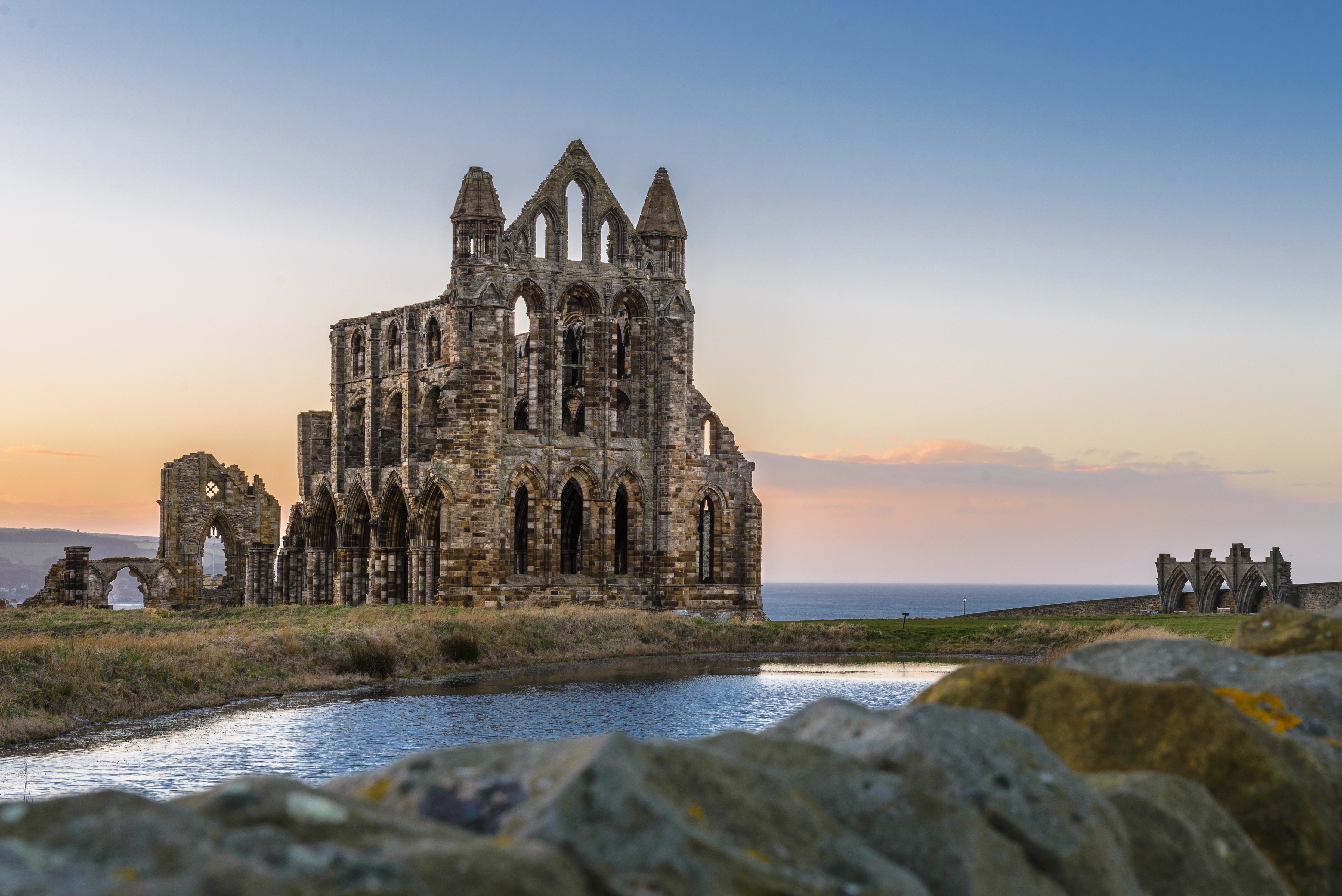 Ruined abbey stands silently beside a small lake, against a serene sunset and distant sea backdrop.