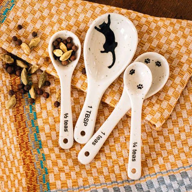 Four ceramic measuring spoons rest on a checkered cloth, adorned with cat silhouettes and paw prints, surrounded by scattered cardamom and peppercorns.