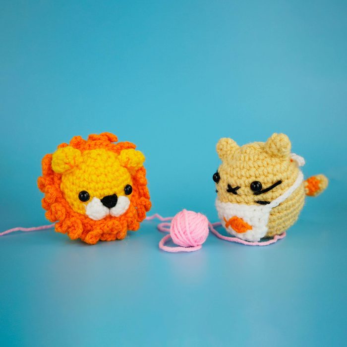 Crocheted lion and cat-like creature sit with pink yarn ball on blue background.