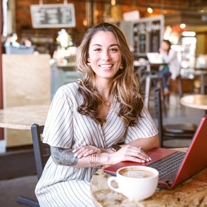 Woman smiling, sitting at a cafÃ© table with a red laptop and latte; people and menu boards visible in the background.