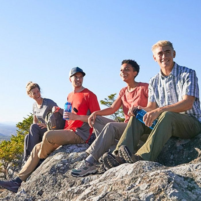 Four people sit on rocks, holding water bottles, enjoying a sunny day outdoors with a clear blue sky and trees in the background.