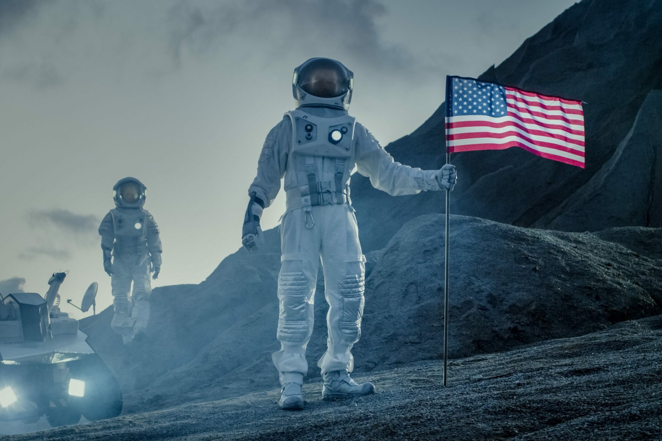 Astronaut holds American flag on rocky lunar surface, with another astronaut and rover in the background under a cloudy sky.